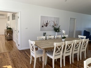 A dining area with a wooden table and white chairs, a painting on the wall, and a plant on the table.