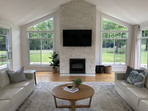 A spacious living room with a stone fireplace, large windows, and a flat-screen TV.