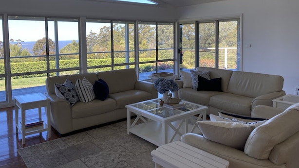 Un salón comedor amplio con ventanas grandes, un sofá color beige, una mesa baja y una pequeña mesa blanca.