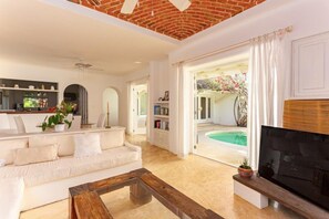 A spacious living room with a white sofa, a coffee table, and a flat-screen TV. There is a pool visible through the open door.