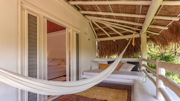 A hammock on a porch with a white railing, a white sofa with cushions, and a thatched roof.