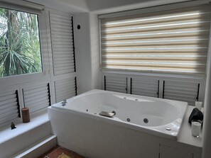 A bathroom with a large bathtub, white walls, and windows with blinds.