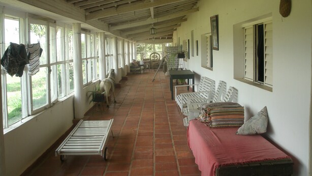 A covered corridor with a long bench, a table with chairs, and a bed with a red blanket.