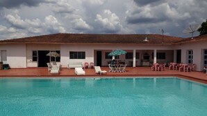 A swimming pool with lounge chairs and umbrellas, a covered seating area, and a building with a tiled roof.