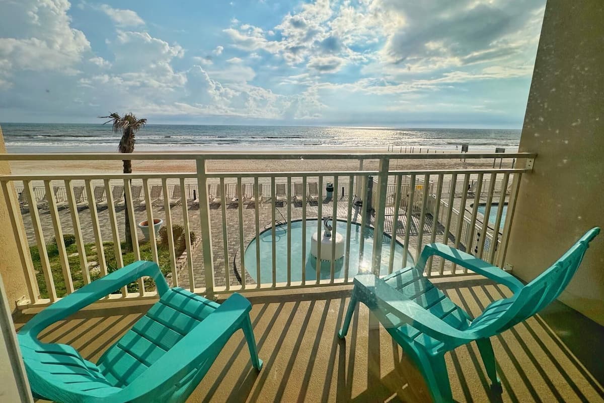 A balcony with a glass table and two turquoise chairs, overlooking a beach with lounge chairs and a palm tree.