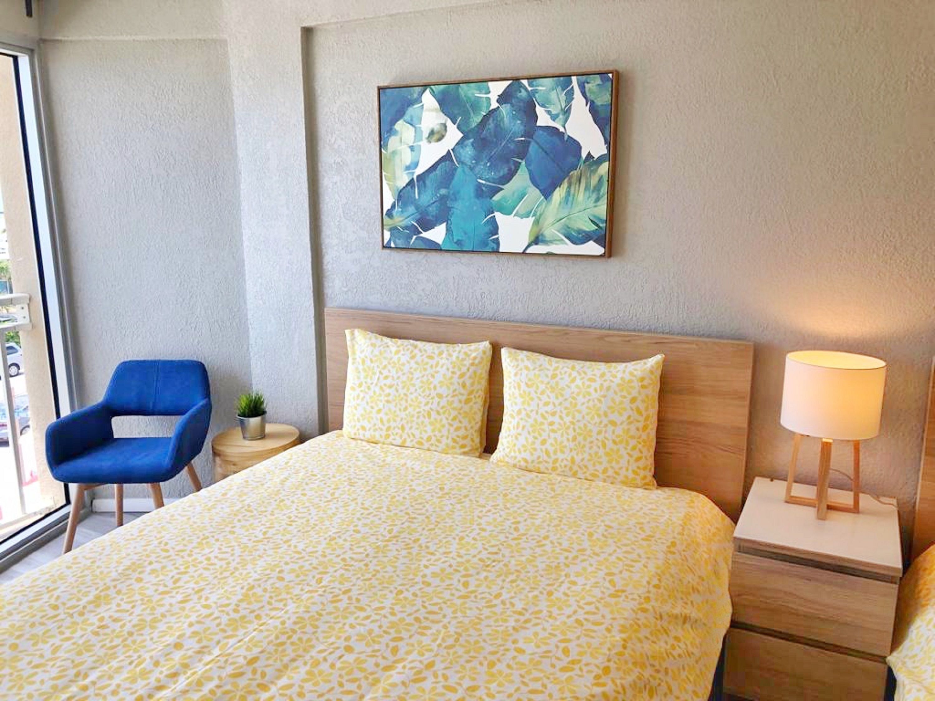 A hotel room with a bed, a blue chair, a nightstand with a lamp, and a painting on the wall.