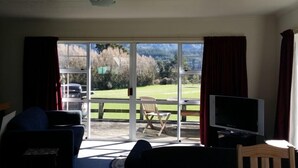 Et værelse med fjernsyn, sofa og et bord med stol. Der er en skydedør til en balkon med udsigt over et grønt landskab, og en bil er parkeret udenfor.
