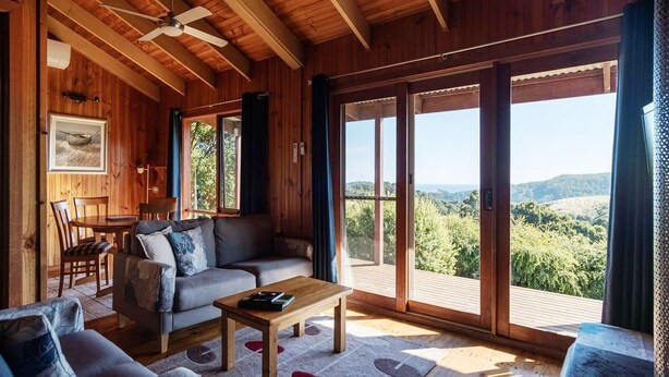 L’intérieur chaleureux d’un chalet, avec un plafond en bois, un canapé, une table basse et de grandes portes-fenêtres coulissantes qui offrent une vue imprenable.