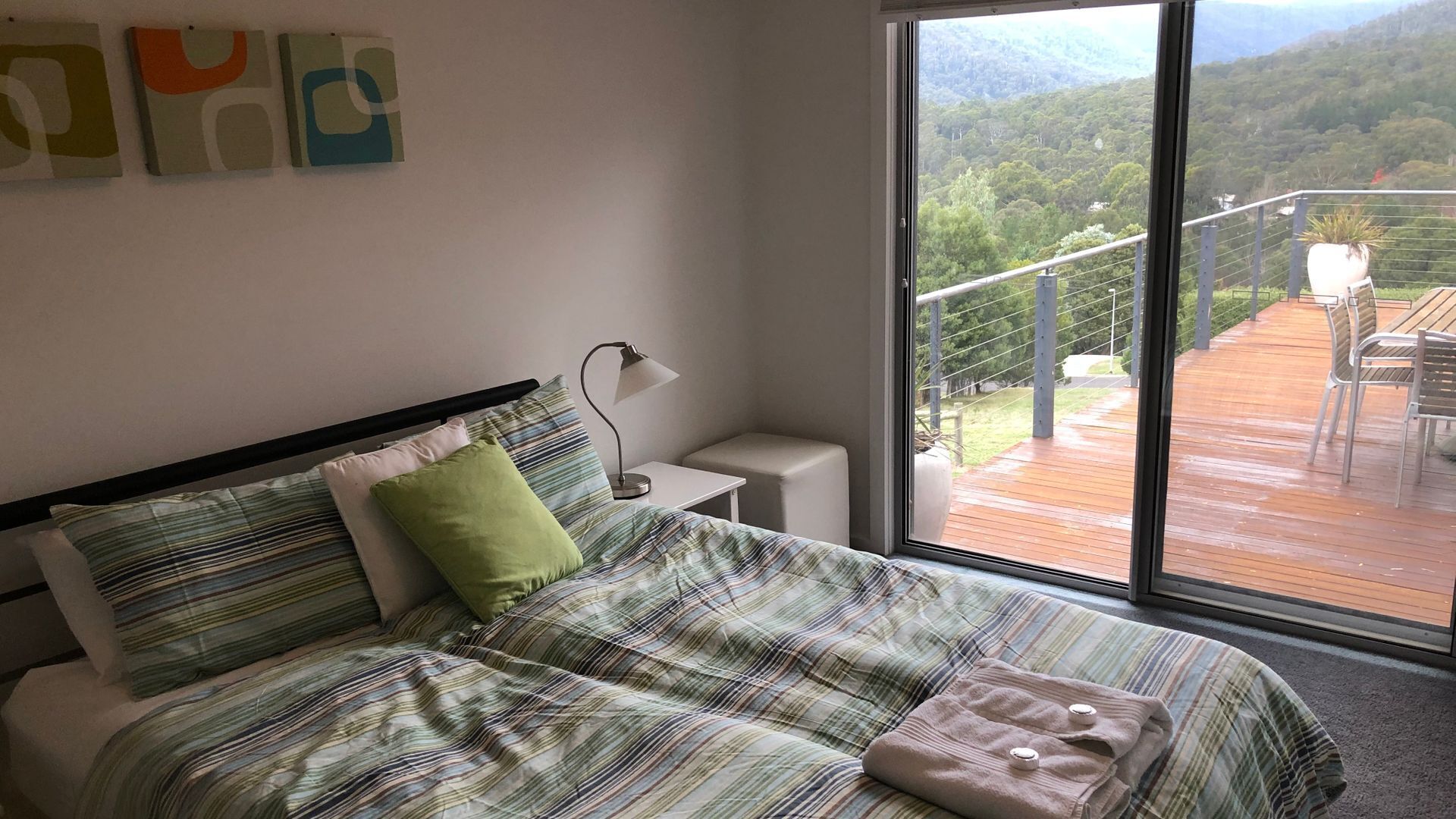 Un dormitorio con cama, almohadas, una mesita de noche con lámpara, y vistas a una terraza y montañas a través de un gran ventanal.
