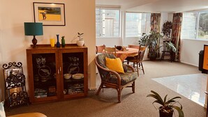Een gezellige woonkamer met een eethoek, met een houten kast met glazen deuren, een rieten stoel met een geel kussen en een tafel gedekt voor vier personen.