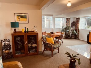 Een gezellige woonkamer met een eethoek, met een houten kast met glazen deuren, een rieten stoel met een geel kussen en een tafel gedekt voor vier personen.