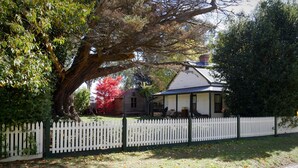 Una casa con un porche, rodeada por una cerca blanca de piquetes y un árbol grande de hojas verdes y rojas.