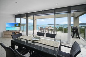 A balcony with a glass table, chairs, a TV, and a grill.