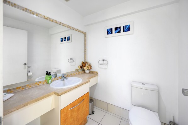 A bathroom with a white sink, mirror, and toilet. There are toiletries on the sink counter and a trash can.