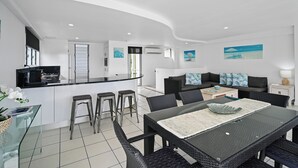 Una cucina moderna con isola centrale, sgabelli da bar, angolo da pranzo con tavolo in vetro e zona living con divano e tavolino.