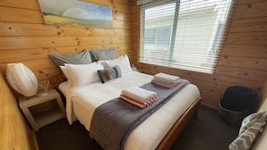 A wooden cabin room with a bed, a nightstand, a window with blinds, and a wall-mounted picture. - Seabed Bach Tokerau Beach (Karikari Peninsula)