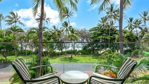 A deck with two striped chairs and a small table, overlooking a lush garden with palm trees and a clear sky.