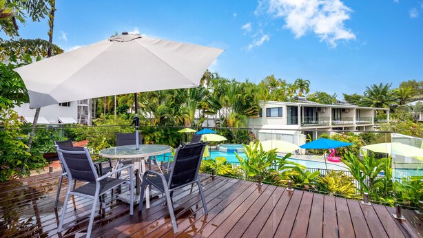 A wooden deck with a round table and chairs, a white umbrella, and a pool area with umbrellas and lounge chairs.