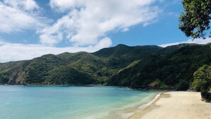 A beach with clear turquoise water, a sandy shore, and lush green hills in the background. - The Oke Beach House (Rawhiti)
