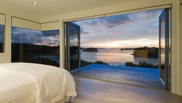 A bedroom with a large bed, a sliding glass door leading to a balcony with a view of a lake and sunset. - The Oke Beach House (Rawhiti)
