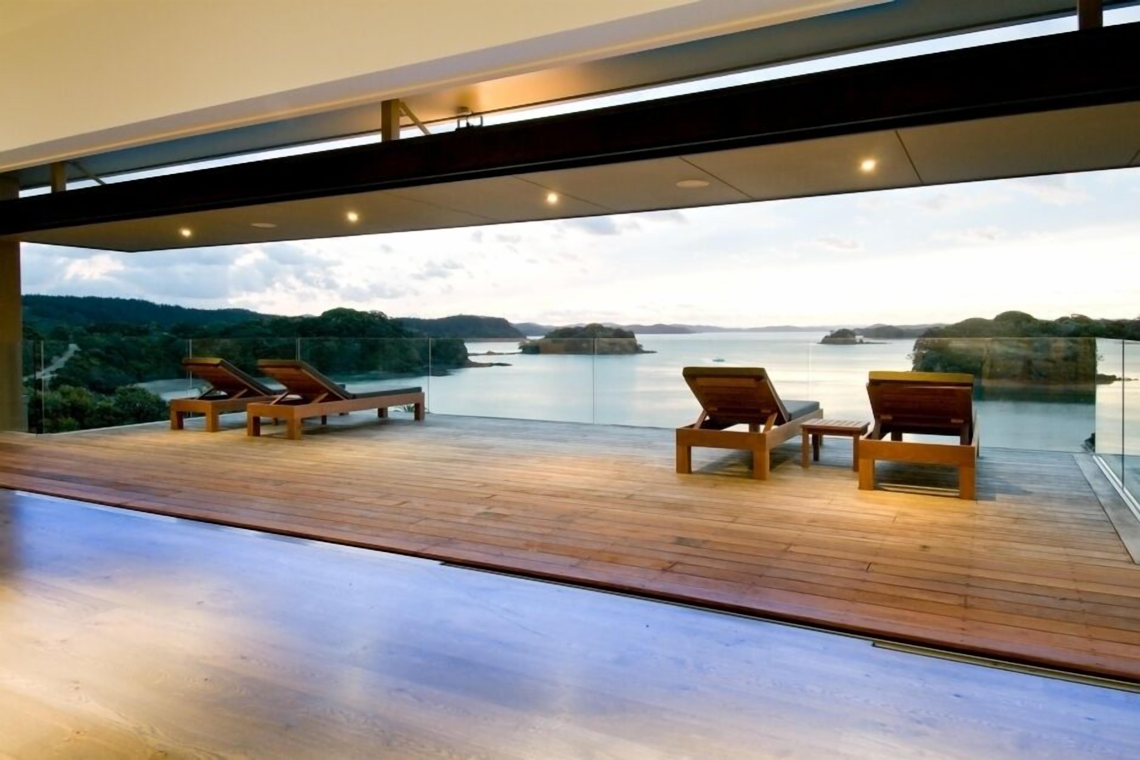 Une terrasse extérieure moderne, dotée d’un plancher en bois et de chaises longues, offrant une vue sur une étendue d’eau paisible et sur des reliefs lointains.