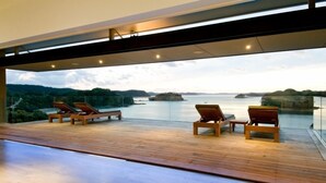 A modern outdoor deck with wooden flooring and lounge chairs, overlooking a calm body of water and distant landforms. - The Oke Beach House (Rawhiti)