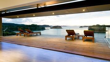 Une terrasse extérieure moderne, dotée d’un plancher en bois et de chaises longues, offrant une vue sur une étendue d’eau paisible et sur des reliefs lointains.