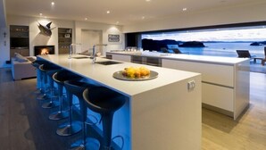 A modern kitchen with a large island, bar stools, and a view of the sea. - The Oke Beach House (Rawhiti)