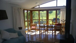 A cozy living room with a sofa, a coffee table, and a rug. There is a dining area with a table and chairs, and a view of a garden through large windows.