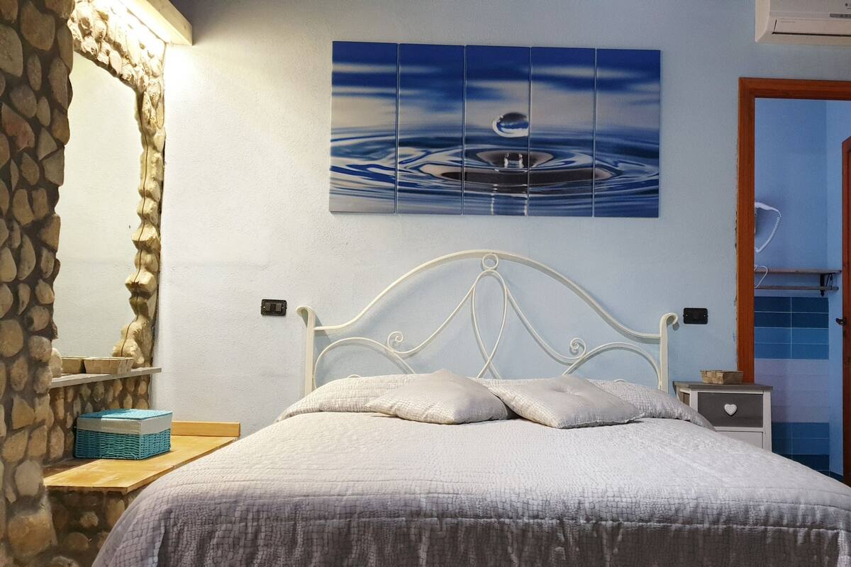 Un dormitorio con pared de piedra, una cama con cabecera blanca, una mesita de noche y un cuadro de una escena acuática.