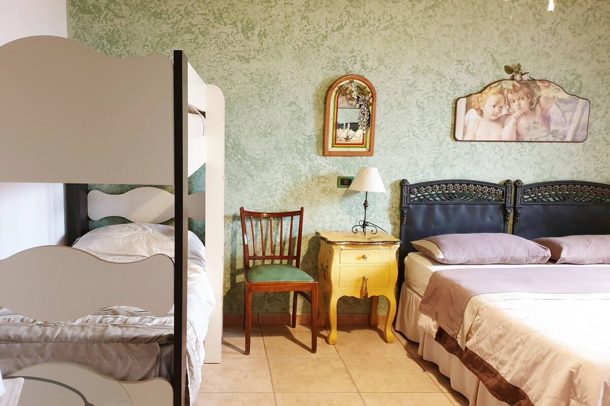 Un dormitorio con una cama litera, una silla, una mesita de noche con lámpara y una cama con almohadas.