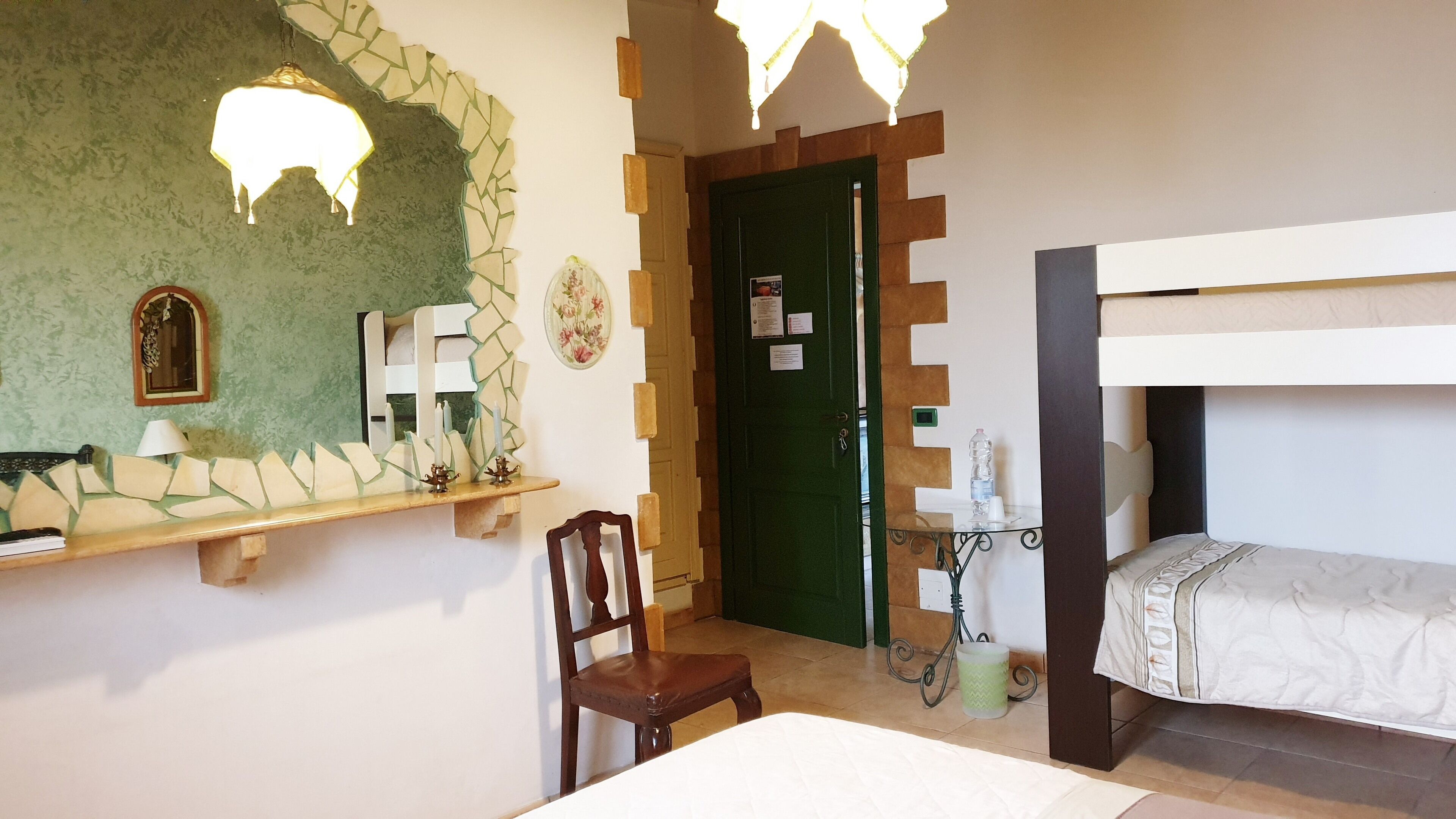 A room with a bed, a chair, a small table with a water bottle, a mirror, and a green door.