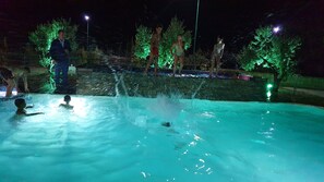 Una piscina nocturna con árboles iluminados y un muro de piedra al fondo.