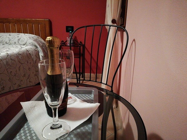 Una habitación con una cama, una silla metálica, una mesa de vidrio y una botella de champán, además de una botella de agua.