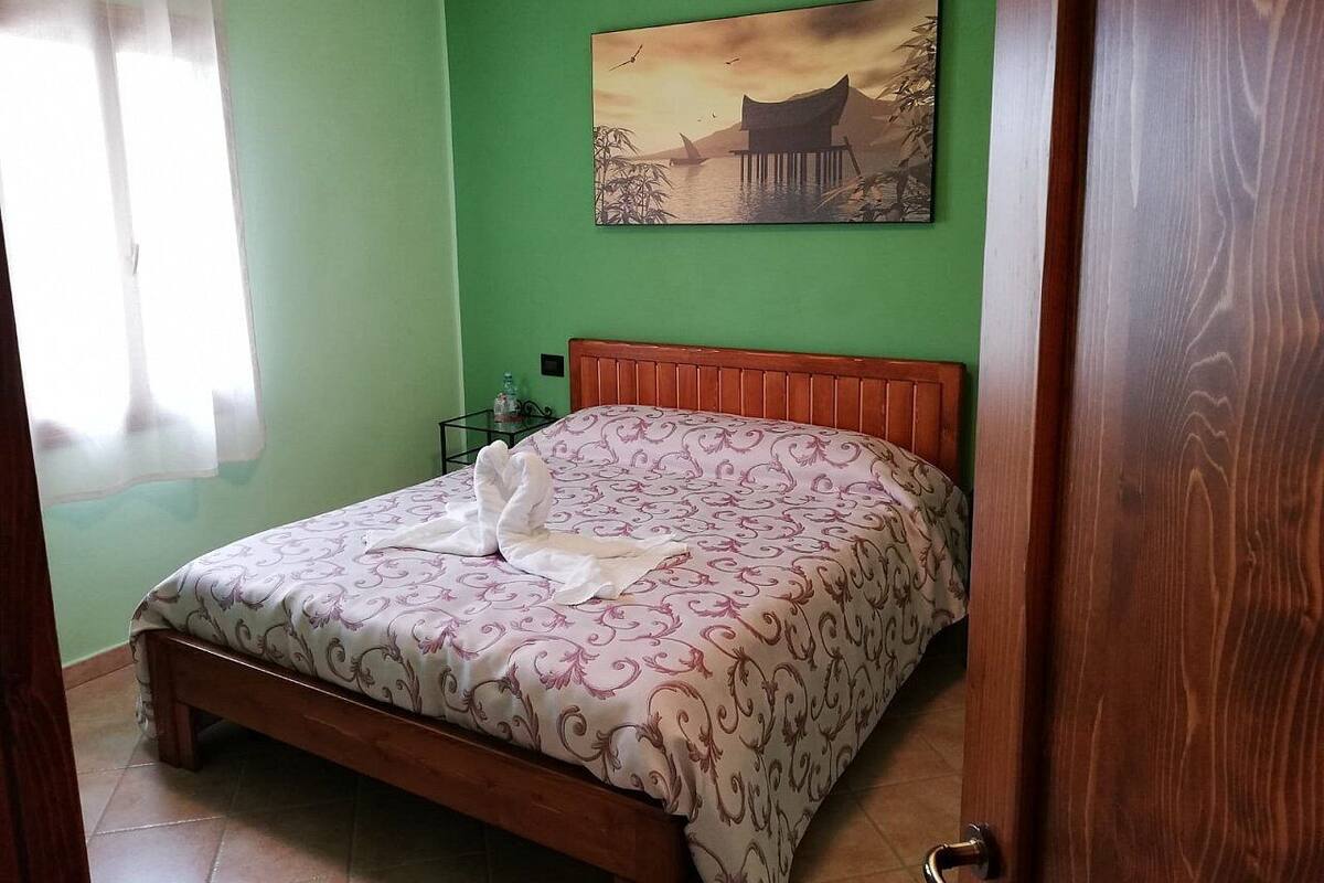 A bedroom with a bed, a nightstand, a painting, and a window with curtains.