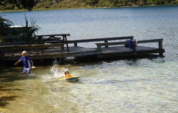 A wooden dock extending into a lake with a child in a boat and another child standing nearby. - Omamari Beach / Kai iwi Lakes / Kauri Forest (Kai Iwi Lakes)