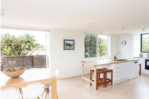 A modern kitchen with a wooden table and chairs, a large window with a view of greenery, and a small island with a sink. - The Peninsula- 4bed Villa in Hastings Point (HASTINGS POINT)