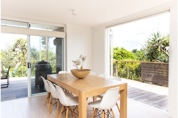 A modern dining area with a wooden table and white chairs, a woven bowl with flowers, and a view of lush greenery through large sliding glass doors. - The Peninsula- 4bed Villa in Hastings Point (HASTINGS POINT)