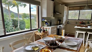 A dining area with a wooden table set for four, surrounded by white chairs. A window offers a view of greenery and hills.