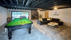 A pool table in a room with a sofa and a small table, large windows, and a fireplace.