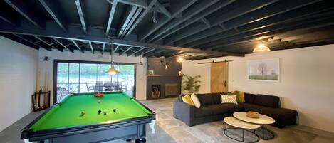 A pool table in a room with a sofa and a small table, large windows, and a fireplace.