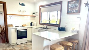 A kitchen with a white countertop, stove, and sink. There are three wooden stools and a window with a view of trees.