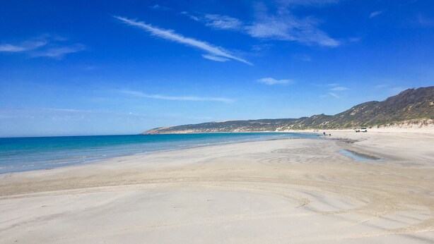 A wide sandy beach with clear blue water and a distant hilly landscape.