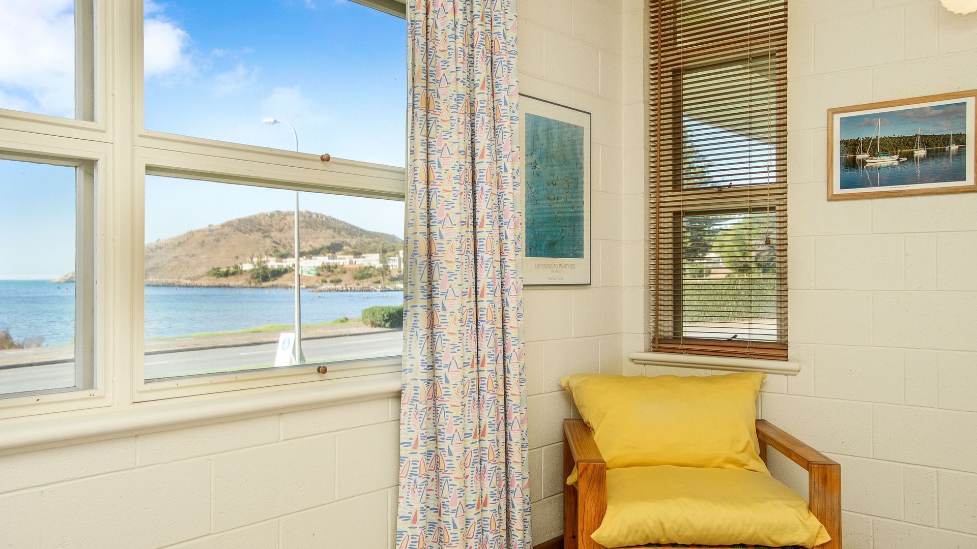 Una habitación con vista al mar, una silla de madera con cojín amarillo y un cuadro titulado "Listening to Paintings" de Barbara Hepworth.
