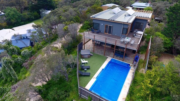 Une maison moderne à deux étages, dotée d’une piscine, d’une terrasse et d’un espace salon extérieur, le tout entouré d’une végétation luxuriante.