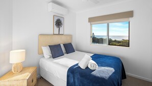 A bedroom with a bed, a nightstand, a lamp, a window with a view, and an air conditioning unit. - 3 Bedroom Penthouse Apartment with a Private Rooftop Spa (Sunshine Coast)