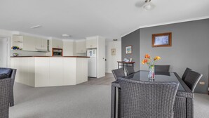 Une cuisine moderne avec des meubles blancs, un îlot central et un coin repas agrémenté de chaises en osier et d’une table en verre.