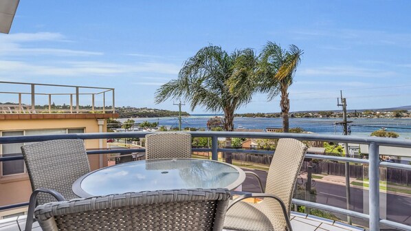 Un balcon doté de fauteuils en osier et d’une table en verre, offrant une vue sur la côte, avec un palmier et une étendue d’eau.