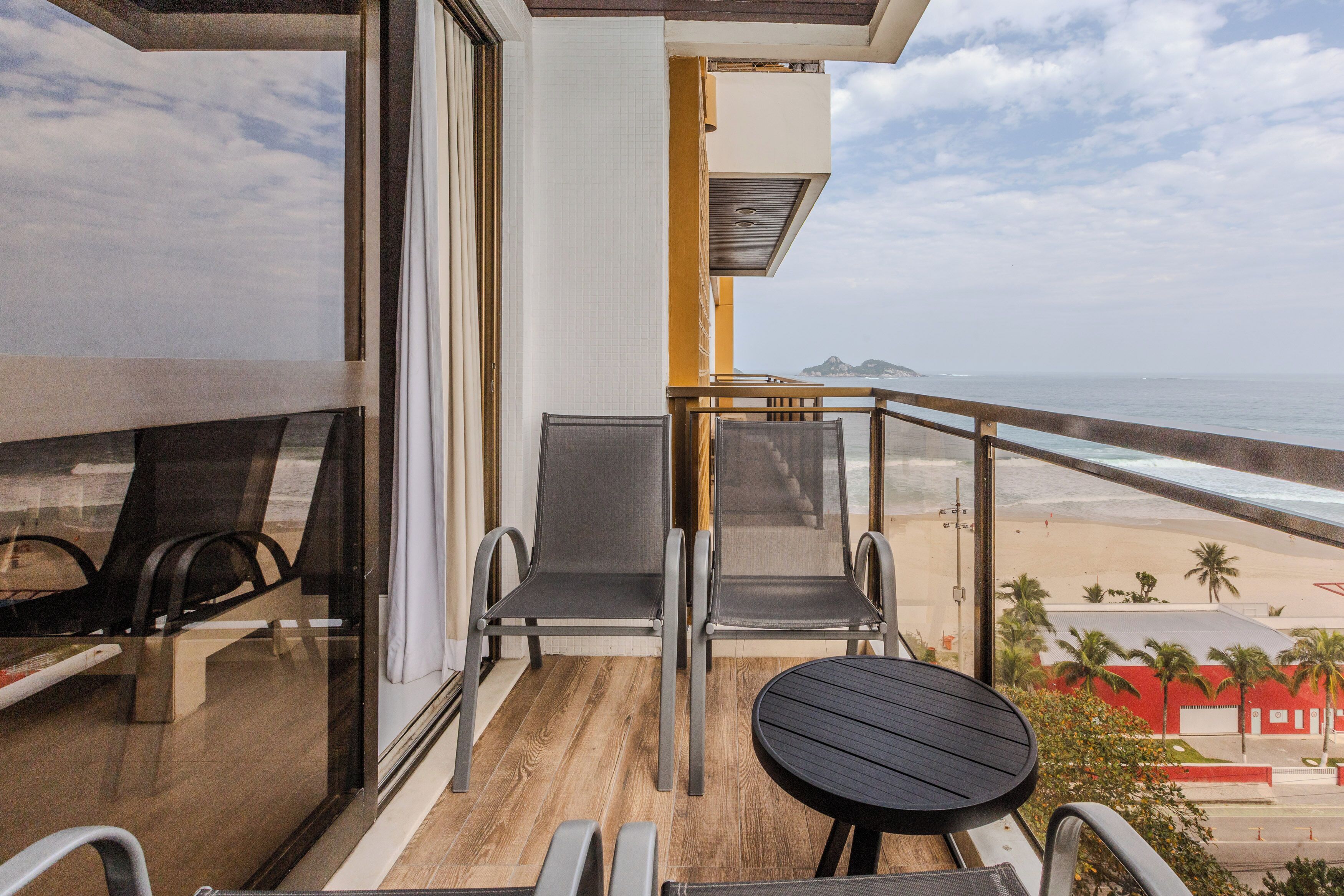 A balcony with two chairs and a small table, overlooking a beach with palm trees and a distant mountain.
