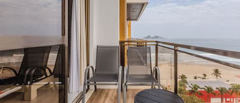 A balcony with two chairs and a small table, overlooking a beach with palm trees and a distant mountain.